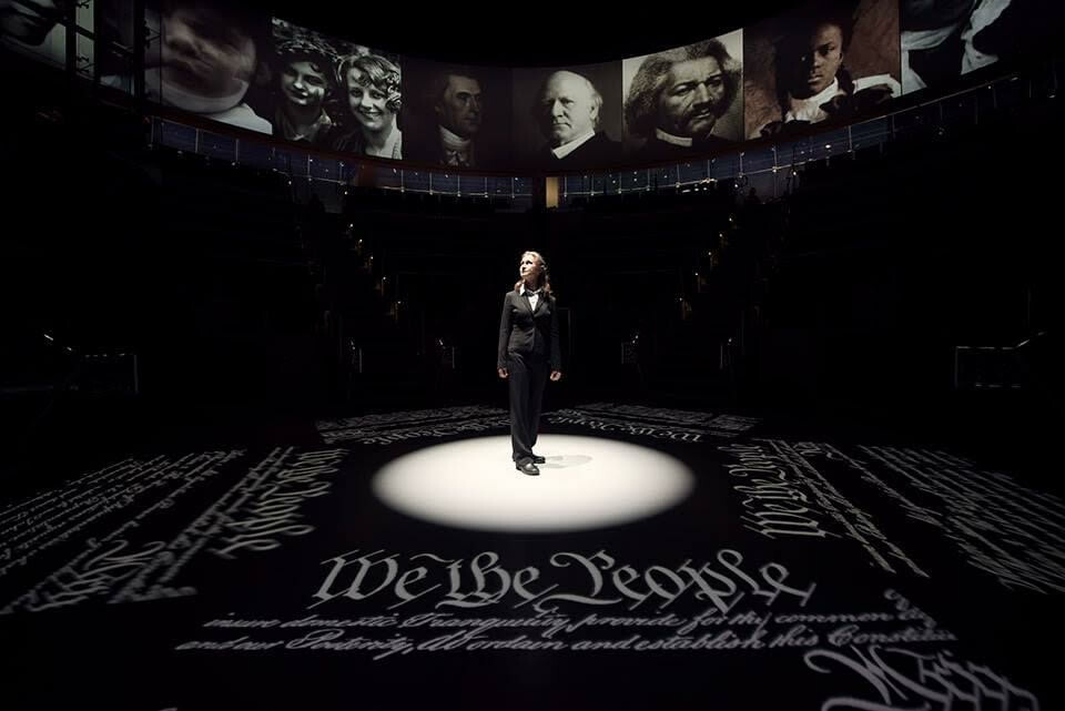 Single person standing on stage in a spotlight, with text from  the Declaration of Independence projected on the floor around her. 