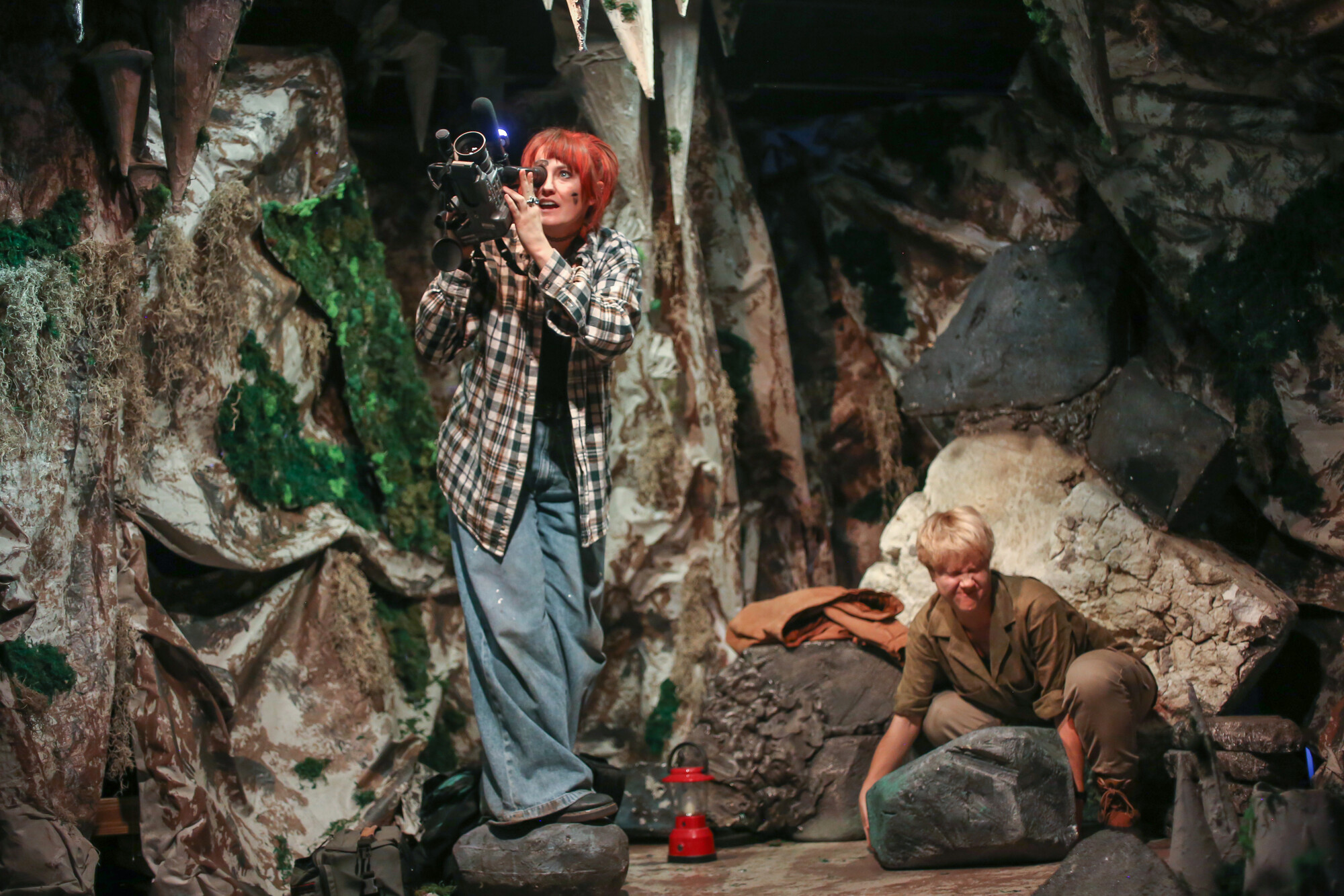 A performer uses a camera while another pushes a prop boulder.