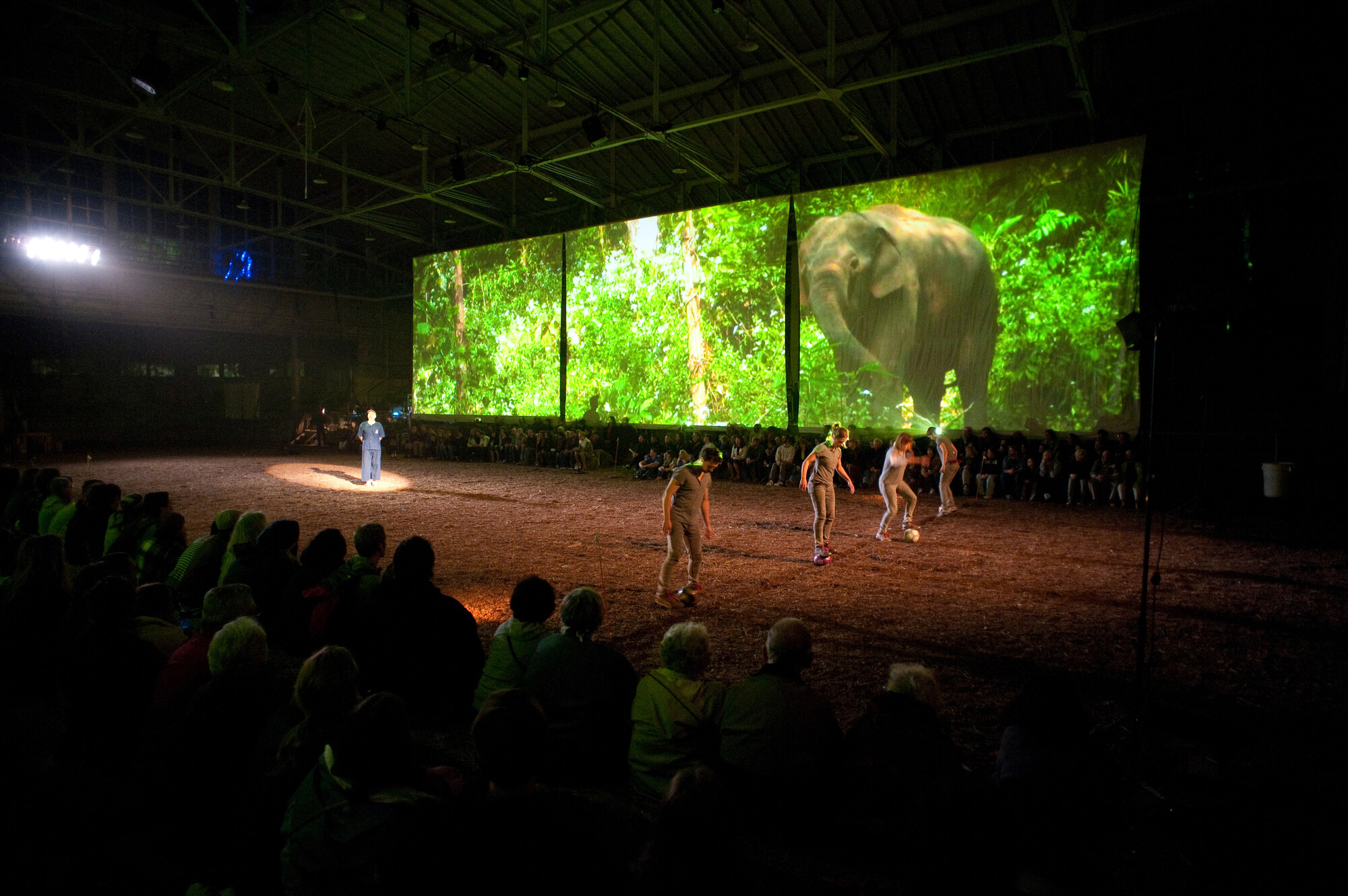 Performers spread out on a sand-covered stage with a screen projecting an image of an elephant in the background.